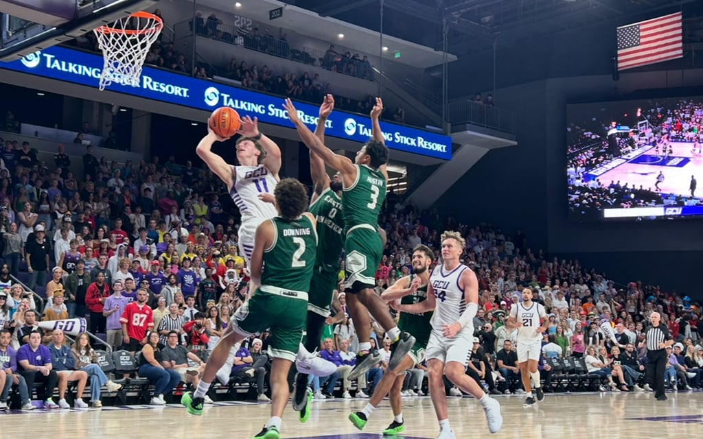 GCU basketball player in white jersey jumping to shoot the ball, with two Eastern New Mexico State players in green jerseys attempting to block, in a crowded arena.