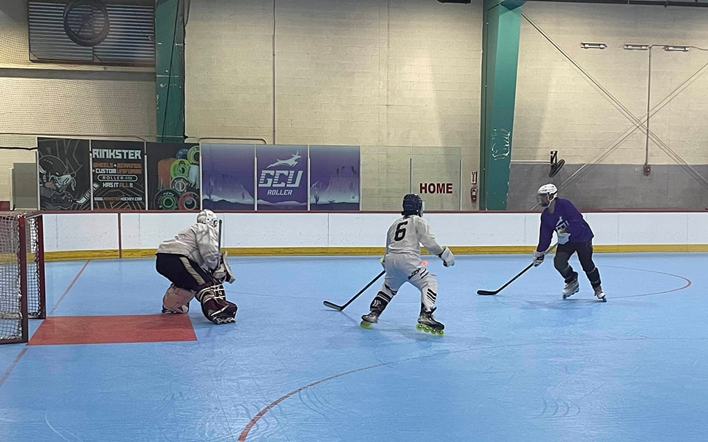 Roller hockey game with three players on a blue rink, including a goalie and two players in motion.