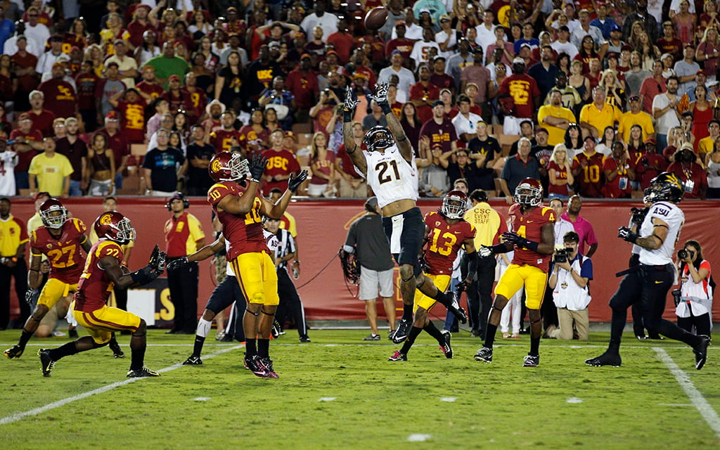 Football game with a player in a white uniform leaping to catch the ball, surrounded by opposing players in red and yellow uniforms.