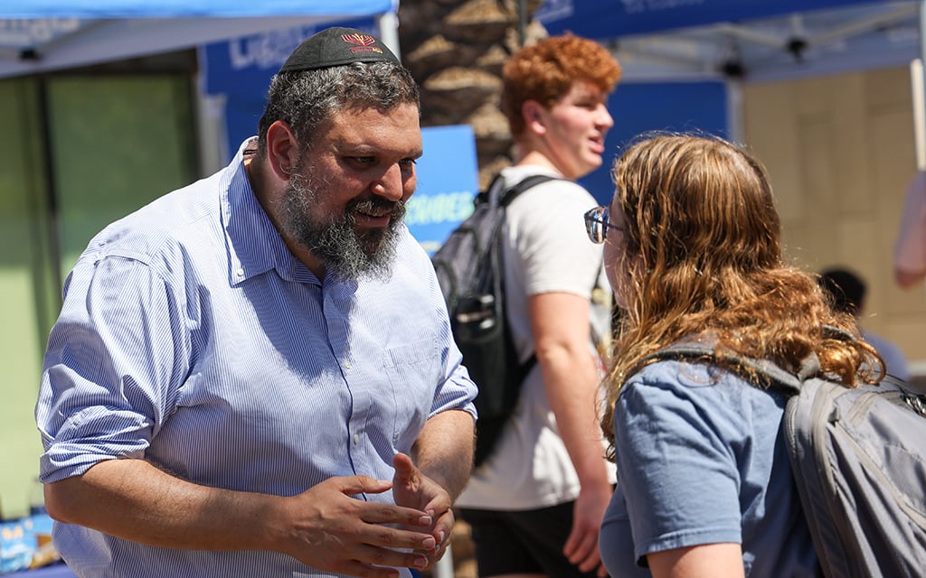 Rabbi Shmuel in a light blue shirt and black kippah engages in conversation outdoors with a woman in glasses and a blue shirt at the "Let There be Light" event.