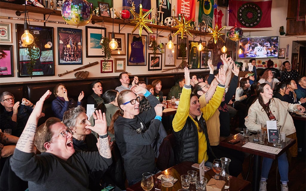 Crowd cheering in a sports bar decorated with sports memorabilia and international flags, watching a televised game.