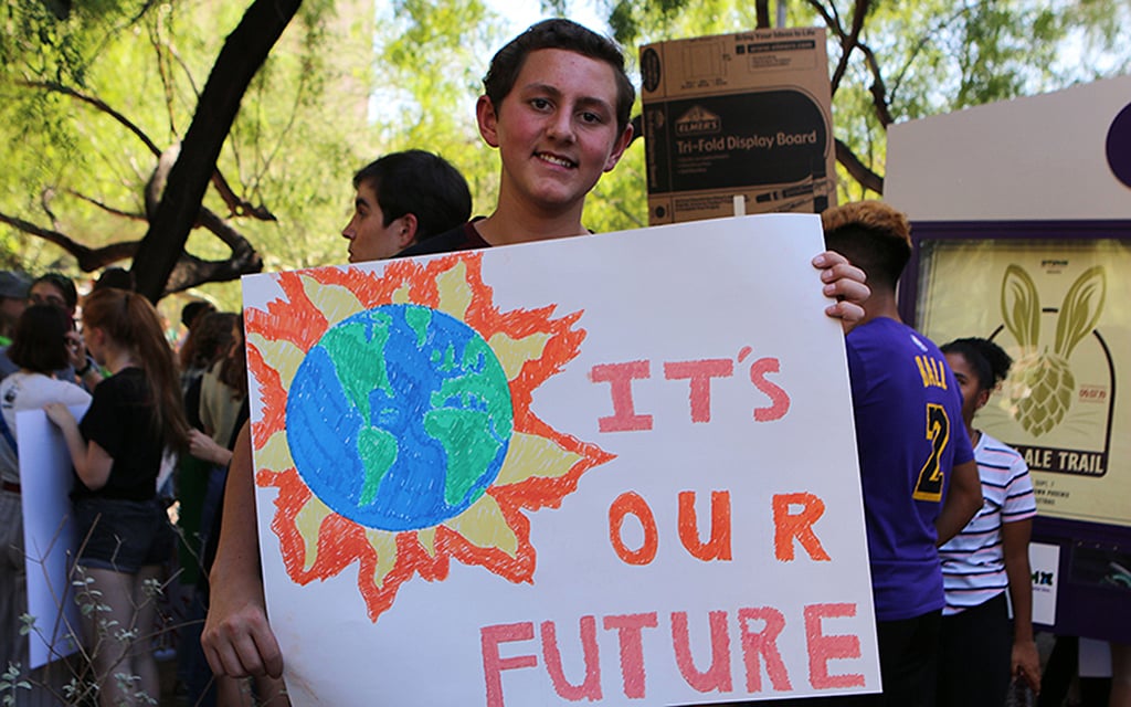 Andrew Bletka holds a sign reading “IT’S OUR FUTURE” with a drawing of the Earth and flames, amidst a crowd in a park.