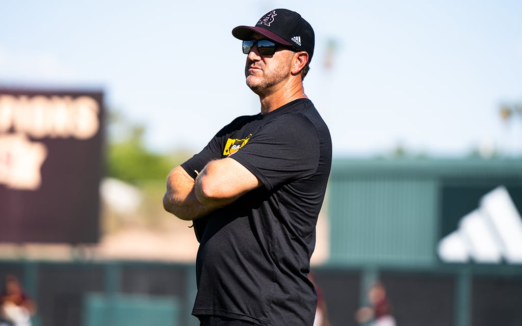 ASU baseball coach Willie Bloomquist wearing a black ASU T-shirt, ASU cap, and sunglasses stands with crossed arms on a sports field.