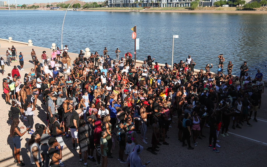 A large crowd gathered on a pathway near Tempe Town Lake with modern buildings in the background.