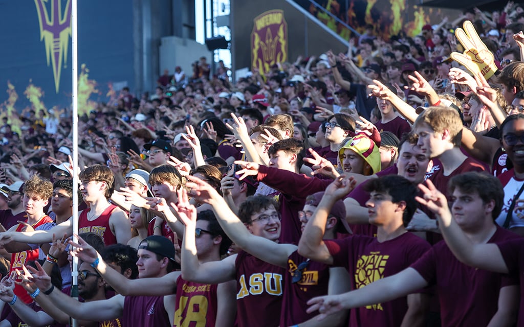 A crowd of ASU football fans in maroon and gold attire, performing the pitchfork gesture in a stadium.