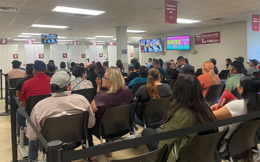 A crowded waiting area with people seated in rows facing numbered service counters and digital screens.