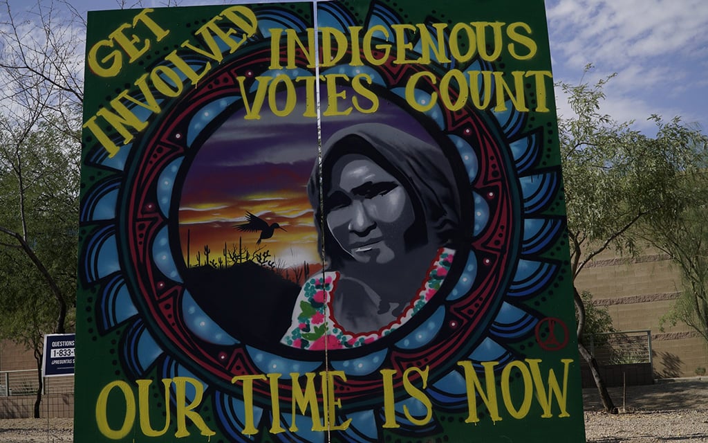 Vibrant mural featuring a grayscale portrait with colorful patterns and sunset scene. Text reads "Get Involved, Indigenous Votes Count, Our Time Is Now."