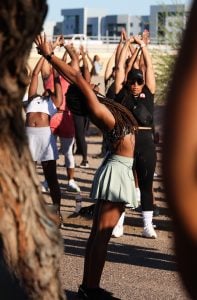 Group of people doing outdoor stretches with arms raised, focus on a woman in a light green skirt.