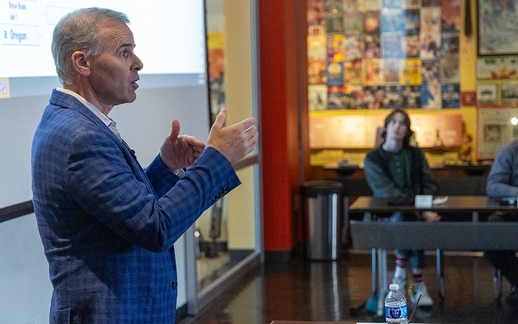 Scott Leightmann, vice president of communications for the Vrbo Fiesta Bowl giving a presentation in a room with a collage-covered wall and audience.