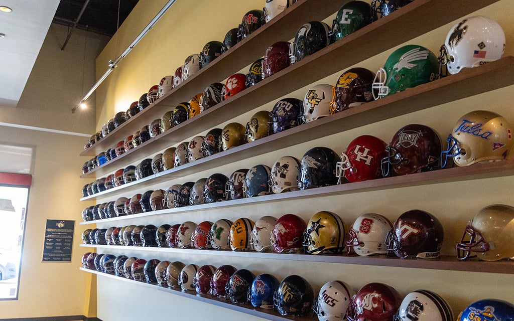 Rowed display of colorful American football helmets on wooden shelves mounted on a yellow wall.