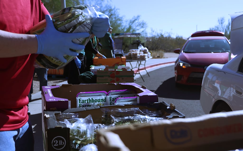 A person wearing blue gloves holds a food package near a table of labeled cardboard boxes. A red car is parked on a sunny driveway.