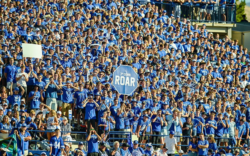 A large crowd of BYU fans in blue and white shirts at LaVell Edwards Stadium, with a sign that says "3rd Down ROAR."