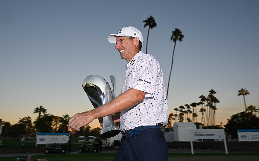 Steven Alker holding a silver trophy on a golf course during sunset, with palm trees in the background.