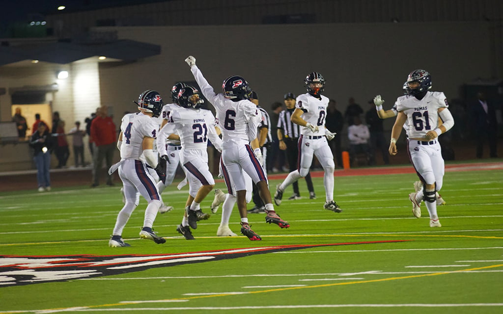 The Perry Pumas’ defense in white uniforms celebrate on a field during a night game.