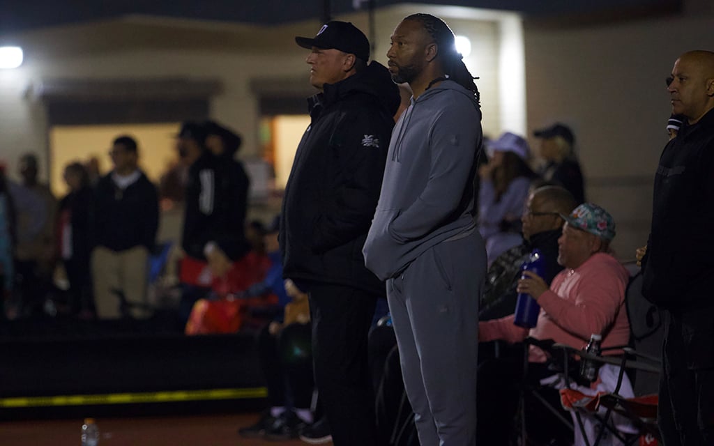 Two men stand outdoors, one in a black jacket and cap, the other being Larry Fitzgerald in a gray tracksuit, surrounded by a blurred crowd.