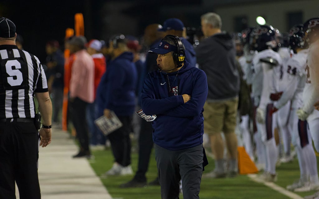 Perry coach Joseph Ortiz on the sideline surrounded by players and a referee during a nighttime game.