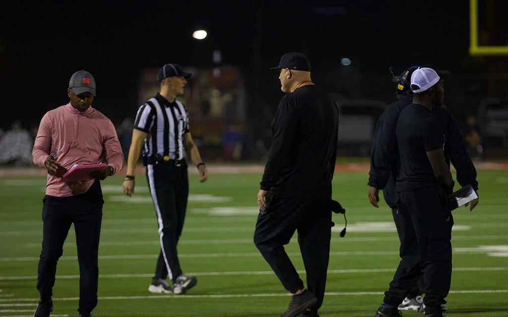 Four men on a football field at night, including one holding a clipboard and a referee walking.