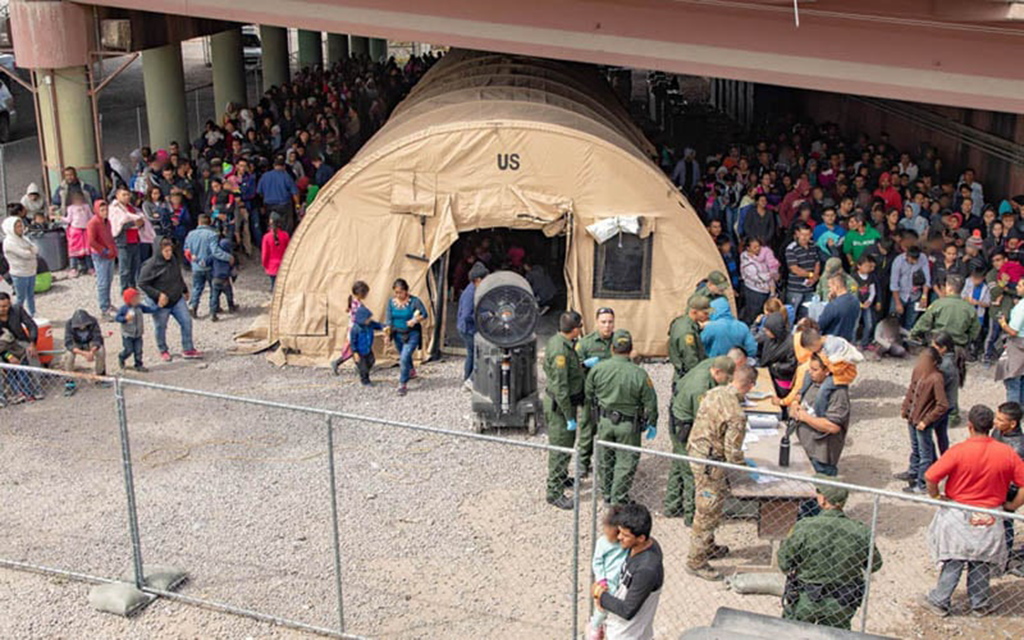 A large crowd lines up outside a tan tent marked "US" under an overpass, with uniformed personnel processing people at a table.