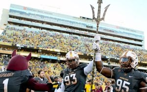 Arizona State University football players and Sparky mascot holding a trident at a stadium.